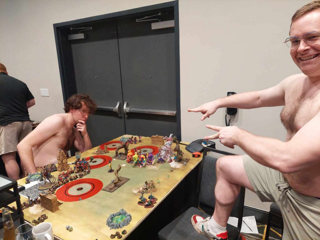 Two men without shirts play Age of Sigmar. One is deep in thought while the other cheers him on.